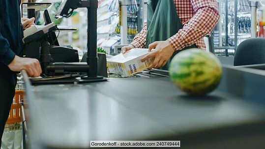 Supermarktkasse mit Melone auf dem Band, im Hintergrund Kassierer und Kunde sowie weitere Artikel