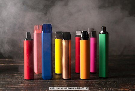 E-Zigaretten in verschiedenen Farben auf Holzuntergrund und vor Rauch