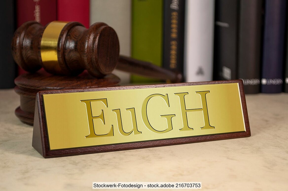 Goldenes Schild mit Aufdruck "EuGH" auf Tisch, im Hintergrund Richterhammer