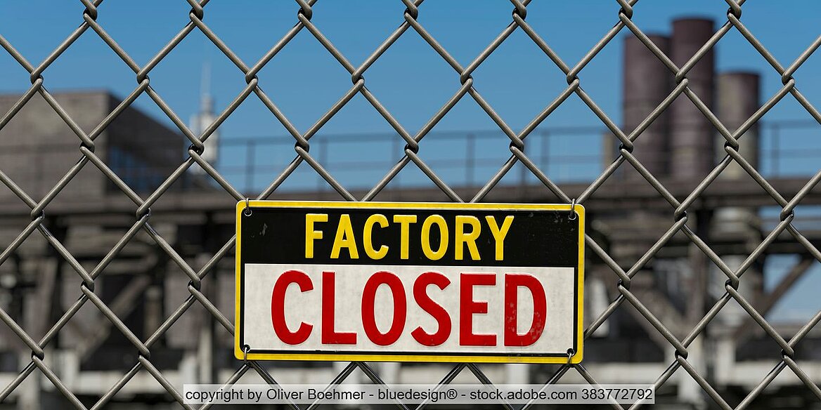 Schild mit Aufschrift "Factory Closed" an Maschendrahtzaun, im Hintergrund Fabrikgebäude und Schornsteine