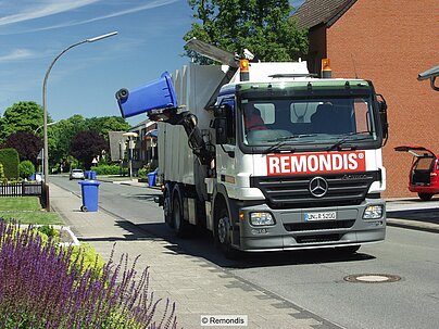 Müllfahrzeug von Remondis bei Leerung von blauen Altpapiertonnen, daneben und im Hintergrund Gärten, Straße, Häuser und Autos 
