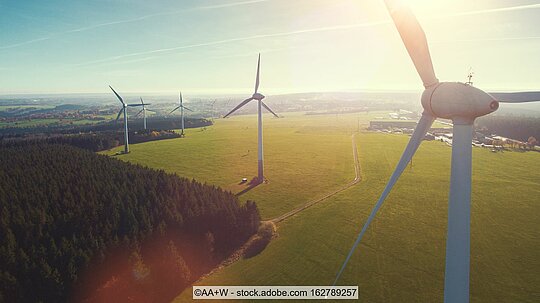 Windkraftanlagen auf grüner Wiese neben Wald und vor blauem Himmel 