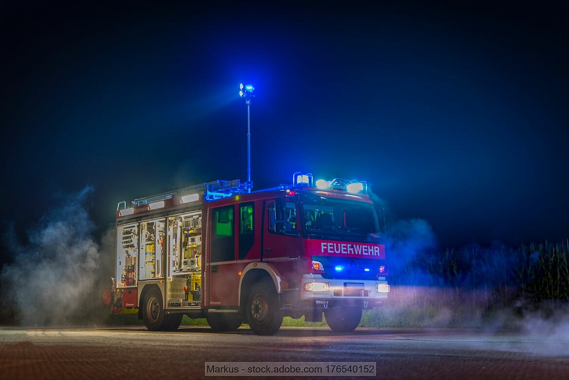 Roter Einsatzwagen der Feuerwehr in der Nacht mit eingeschaltetem Blaulicht auf Platz mit blau leuchtender Laterne
