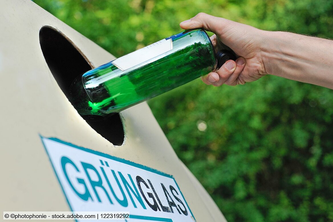 Eine Hand wirft eine grüne Flasch in einen Altglascontainer.