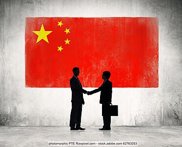 Symbolbild: Silhouette von zwei Männern in Business-Anzügen, die sich die Hand schütteln. Im Hintergrund die chinesische Flagge.