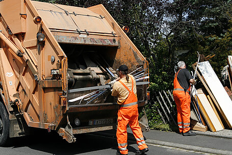 Zwei Müllwerker laden Sperrmüll in einem Müllwagen ab.