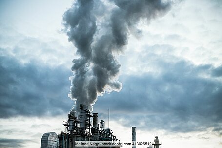 Grauer Rauch tritt aus Schornsteinen von Industrieanlage aus, im Hintergrund bewölkter Himmel