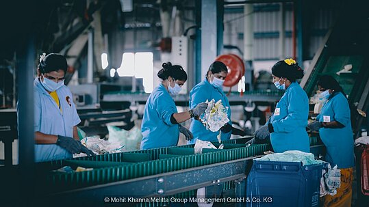 Arbeiterinnen in blauen Arbeitskitteln sortieren Abfälle an Laufband aus