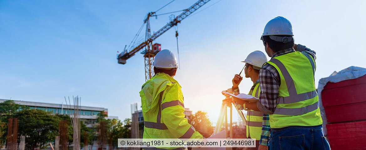 Drei Männer mit gelben Warnwesten stehen auf einer Baustelle und schauen hoch zu einem Kran.
