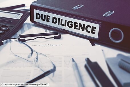 Ordner mit Aufschrift "Due Dilligence" auf Schreibtisch neben Stiften, Zetteln, einer Brille owie einem Taschenrechner