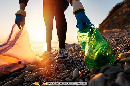 Person sammelt Plastikflasche und weiteren Müll an Küste von Meer oder Fluss, im Hintergrund Sonne