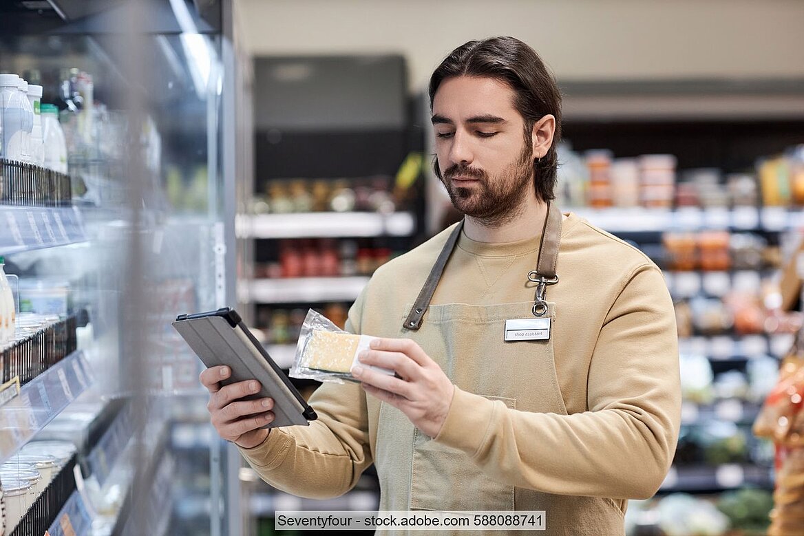 Supermarktmitarbeiter in Schürze vor Regal mit verschweißter Käsepackung in der einen und Tablet in der anderen Hand