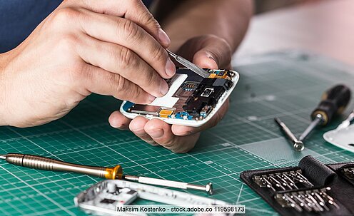 Hände bei der Reparatur von aufgeschraubtem Smartphone, im Vordergrund Werkzeug