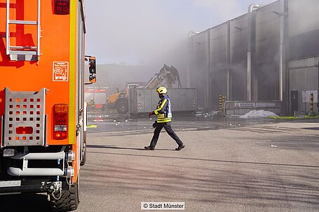 Ein Feuerwehrmann läuft vor einem Einsatzfahrzeug entlang und im Hintergrund raucht ein Gebäude.