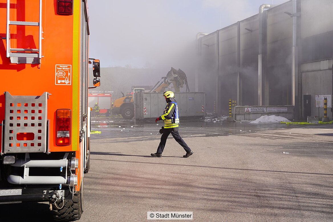Ein Feuerwehrmann läuft vor einem Einsatzfahrzeug entlang und im Hintergrund raucht ein Gebäude.