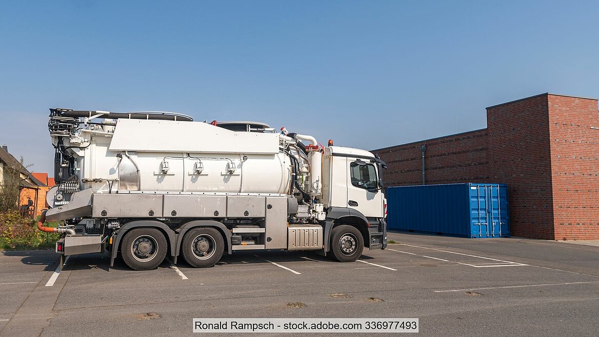 Lkw mit Saug- und Pumptechnik sowie Tank als Aufbauten auf grauer Betonfläche vor brauner Backsteinwand und blauem Container