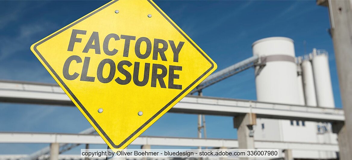 Gelbes Schild mit Aufdruck "FACTORY CLOSURE" in schwarzer Schrift, im Hintergrund Industrieanlagen und Türme 