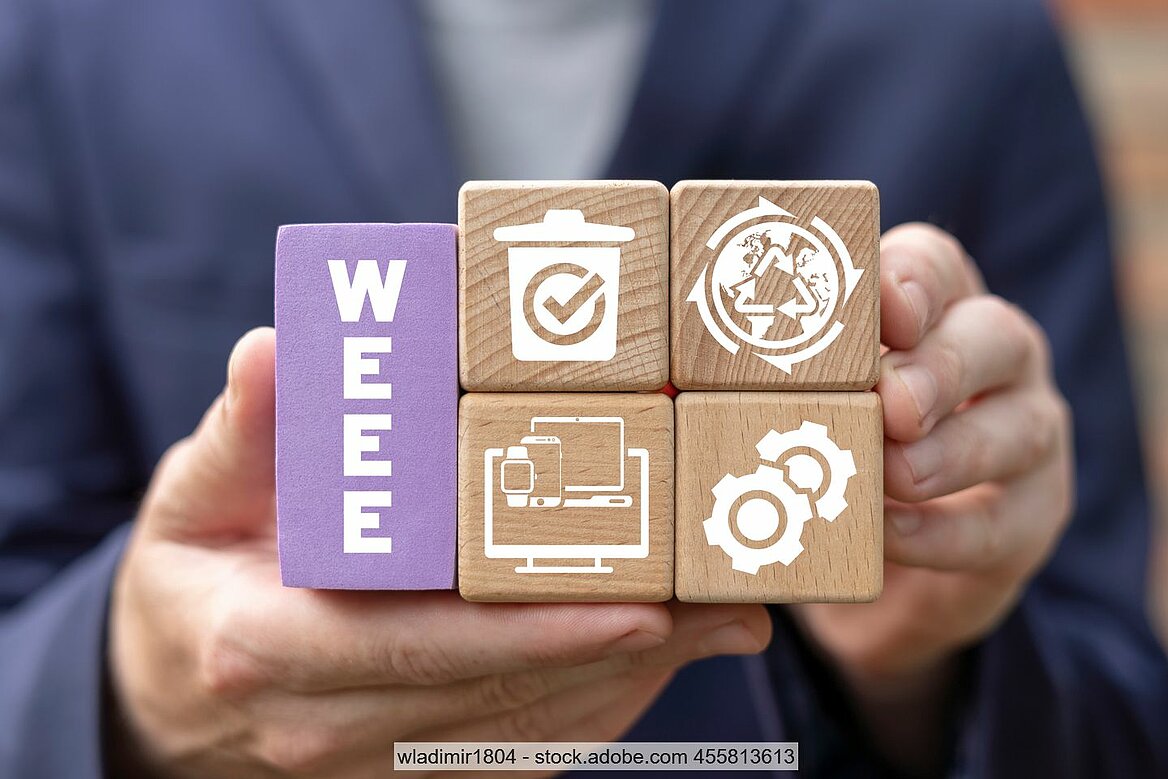 Hand mit Holzsteinen, die mit "WEEE" und verschiedenen Symbolen zu Elektrogeräten, Mülltonnen und Kreislaufsymbolen bedruckt sind