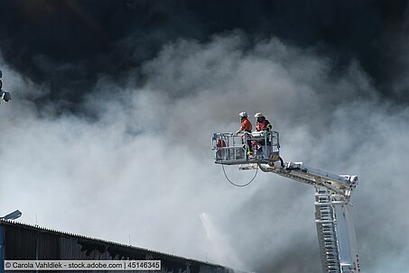 Zwei Feuerwehrmänner stehen auf einer Drehleiter über einem rauchenden Gebäude.