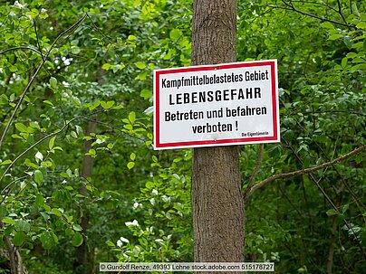 Weißes Schild mit rotem Rand und schwarzer Schrift, die vor Lebensgefahr wegen Kampfmittelbelastung warnt, an Baumstamm im Wald