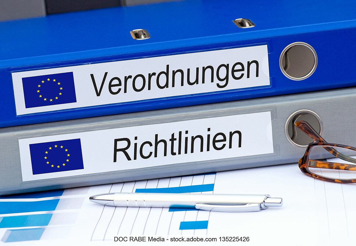 Blauer und grauer Ordner mit EU-Fahne und "Verordnungen" bzw. "Richtlinien" als Aufdruck, davor liegen Stift und Brille auf Papier