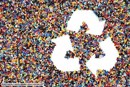 Recyclingsymbol mit drei Pfeilen vor Hintergrund aus bunten Kunststoffpellets.