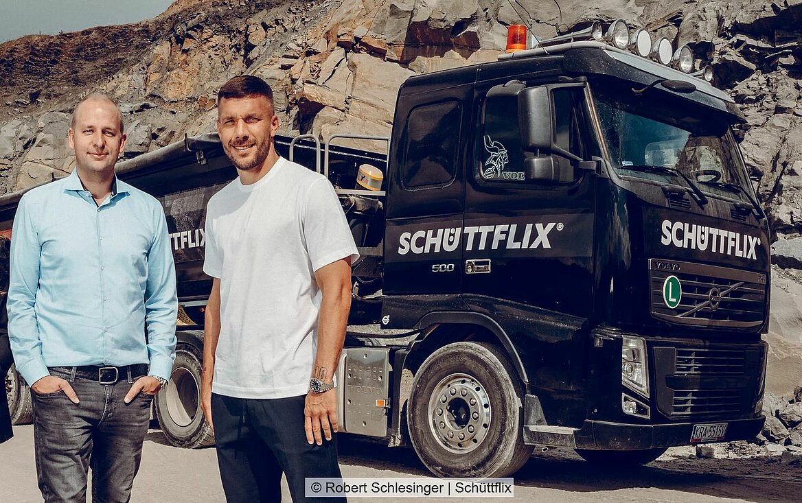 Lukas Podolski steht mit einem Mann vor einem LKW des Logistikunternehmens Schüttflix.