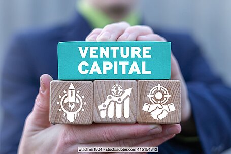 Hand hält Holzwürfel mit Aufschrift "Venture Capital" und Symbolen für aufsteigende Rakete, Diagramm mit aufsteigenden Balken und Handschlag