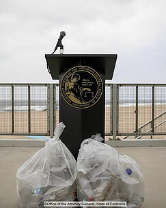 Ein Redepult mit dem Wappen des Generalstaatsanwalts von Kalifornien, auf einer Art Standpromenade stehend. Vor dem Pult liegen zwei große transparente Müllbeutel, die mit Kunststoffabfällen gefüllt sind.