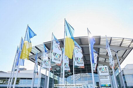 Flaggen der IFAT und die Nationalflagge der Ukraine hängen vor einem Gebäude der Messe München.