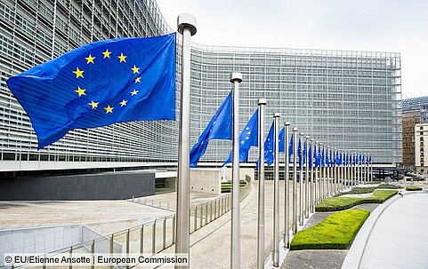 Foto des Gebäudes der EU-Kommission in Brüssel mit einer Reihe von Fahnenmasten mit der EU-Flagge im Vordergrund.