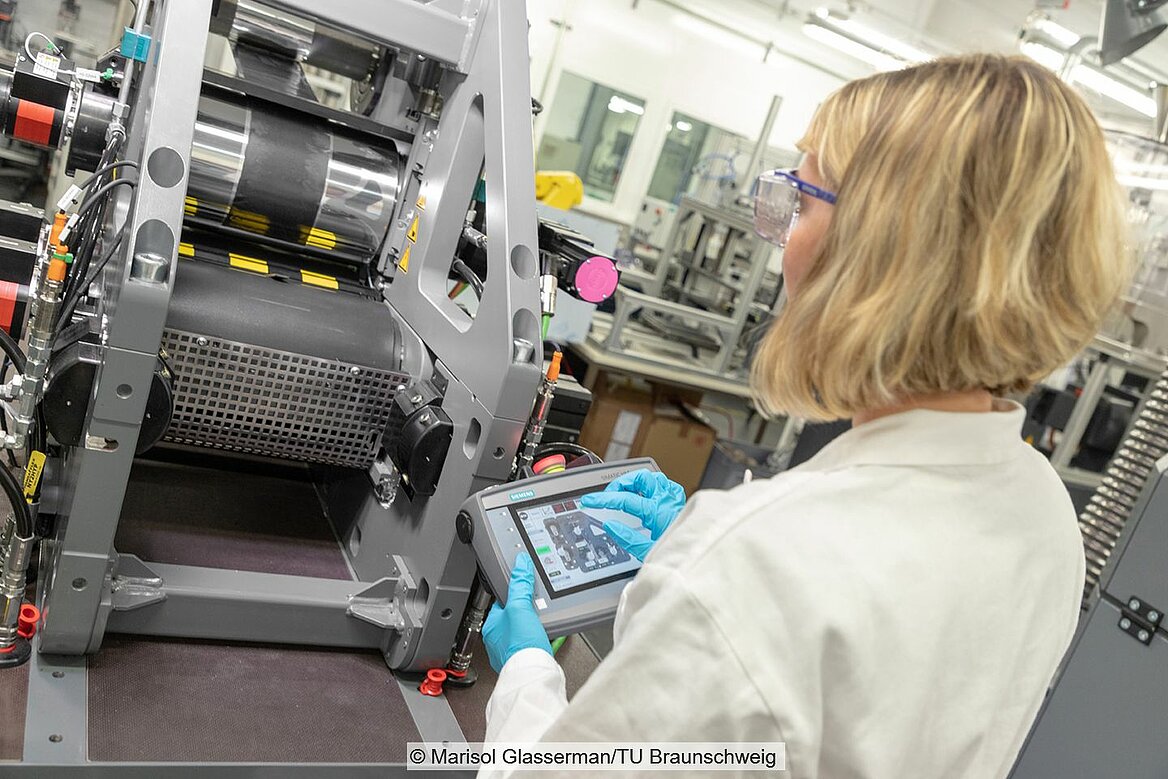 Frau in weißem Laborkittel steht in Halle mit Maschinen und hält Gerät mit Bildschirm in der Hand