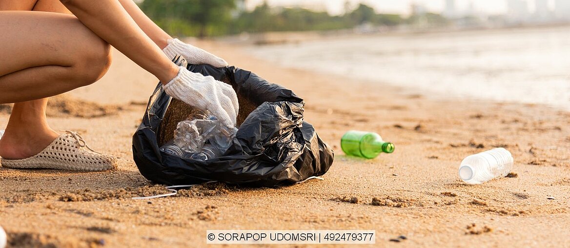 Flaschen liegen an einem Strand und werden in einem schwarzen müllsack gesammelt.