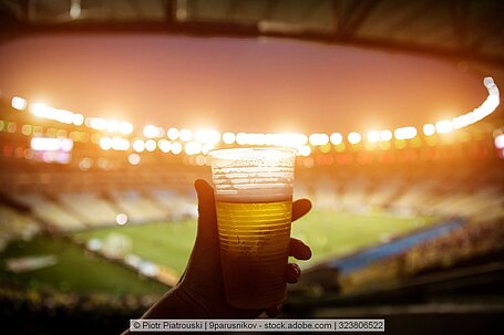 Ein Einwegbecher gefüllt mit Bier wird in einem Stadion ins Licht gehalten.