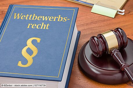 Dickes blaues Buch mit goldenem Aufdruck "Wettbewerbsrecht &" daneben Richterhammer, beides auf Holztisch"