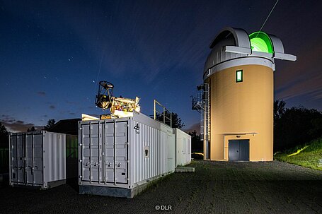 Turm des Johannnes-Kepler-Observatoriums mit geöffnetem Dach aus dem grüner Lichtstrahl des Teleskops leuchtet neben grauen Containern und vor Bäumen sowie sternenklarer Nacht