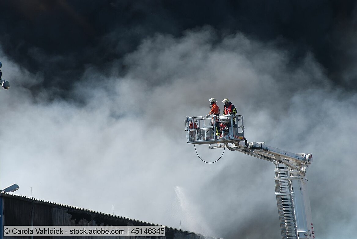 Zwei Feuerwehrmänner stehen auf einer Einsatzleiter. Dichter Rauch steigt aus dem Gebäude auf.