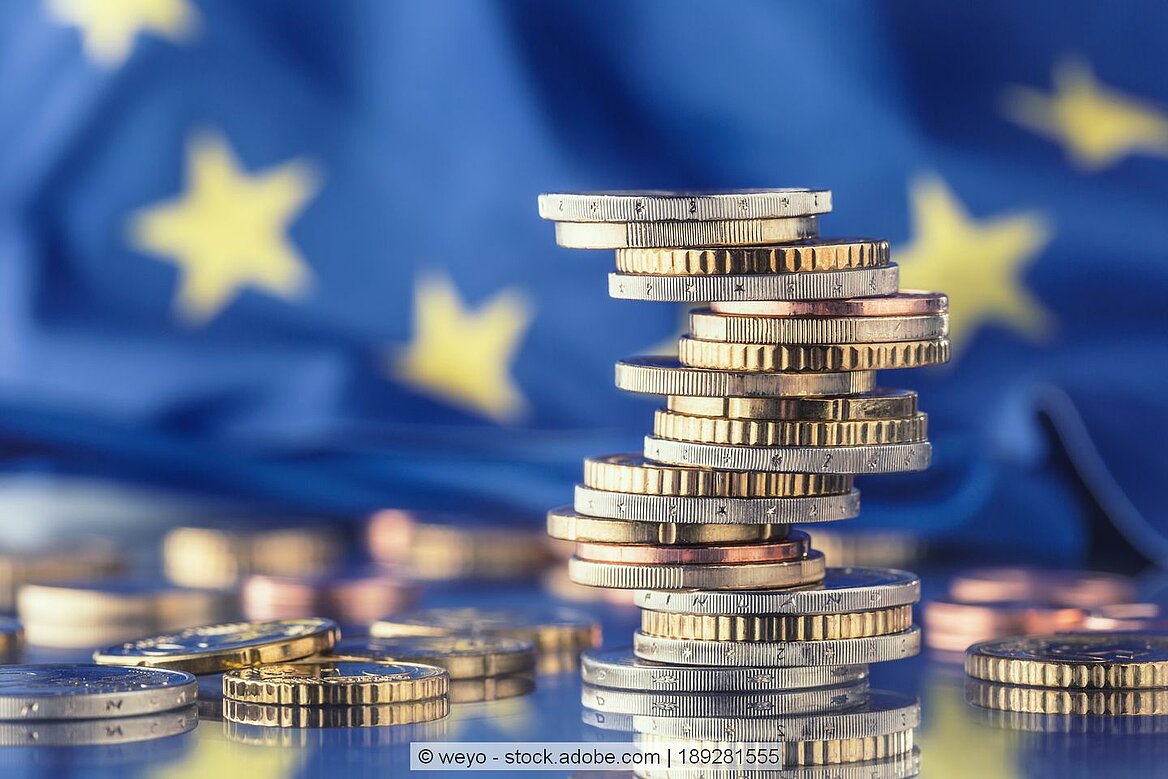 Euro-Münzen sind aufeinander gestapelt und liegen vor einer EU-Flagge.