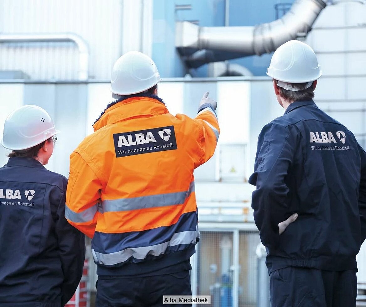 Rückansicht von Alba-Mitarbeitern, die blaue und orange-farbene Jacken mit Alba-Logo tragen und auf Industrieanlage schauen