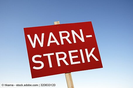 Rotes Schild mit Aufschrift "Warnstreik" vor hellblauem Hintergrund