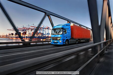 Lkw mit Container fährt über eine Brücke, im Hintergrund sind Hafenanlagen zu sehen. 