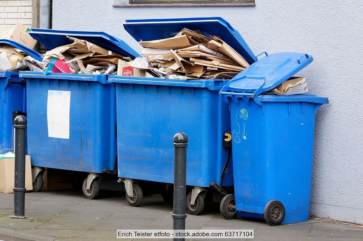 Hoch mit Altpapier gefüllte blaue Müllgroßbehälter und kleinere Tonnen vor einer Hauswand. 