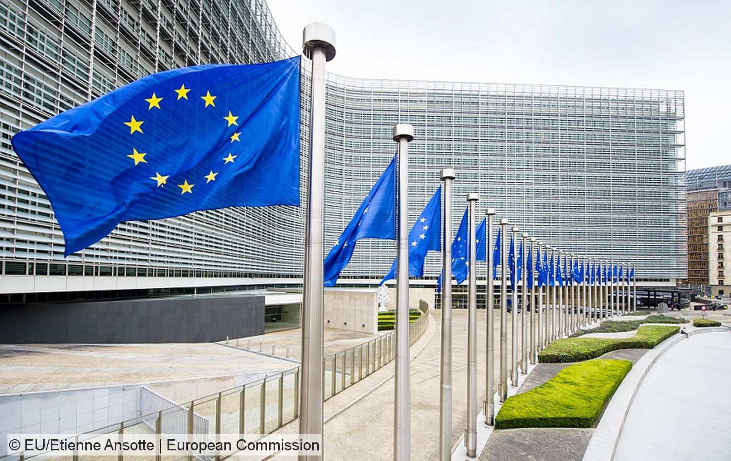 Foto des Gebäudes der EU-Kommission in Brüssel mit einer Reihe von Fahnenmasten mit der EU-Flagge im Vordergrund.