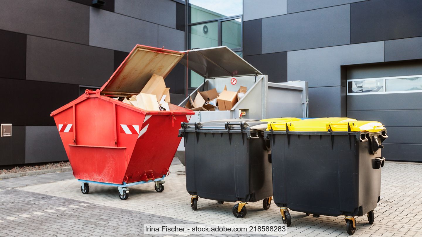 Roter Abfallcontainer und zwei große Mülltonnen mit gelbem Deckel vor Industriehalle und Papierpresse