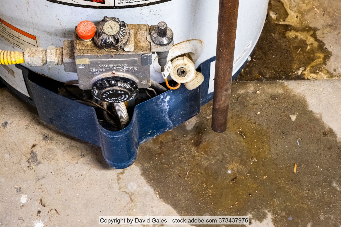 Unterer Teil eines alten Boilers mit verdreckten Reglern, Rohren und Kabeln auf teilweise feuchtem Untergrund