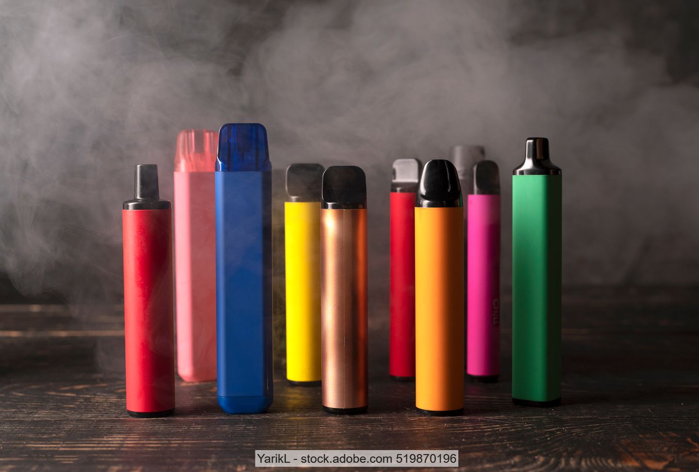 E-Zigaretten in verschiedenen Farben und Größen, teilweise umhüllt von Rauch auf Holzuntergrund