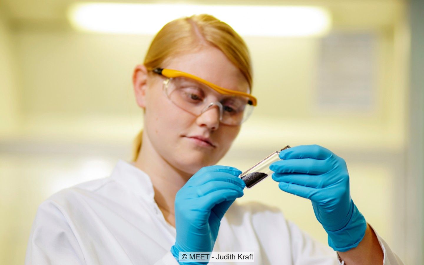 Eine Forscherin im weißen Kittel und mit handschuhen hält ein Reagenzglas in der Hand.