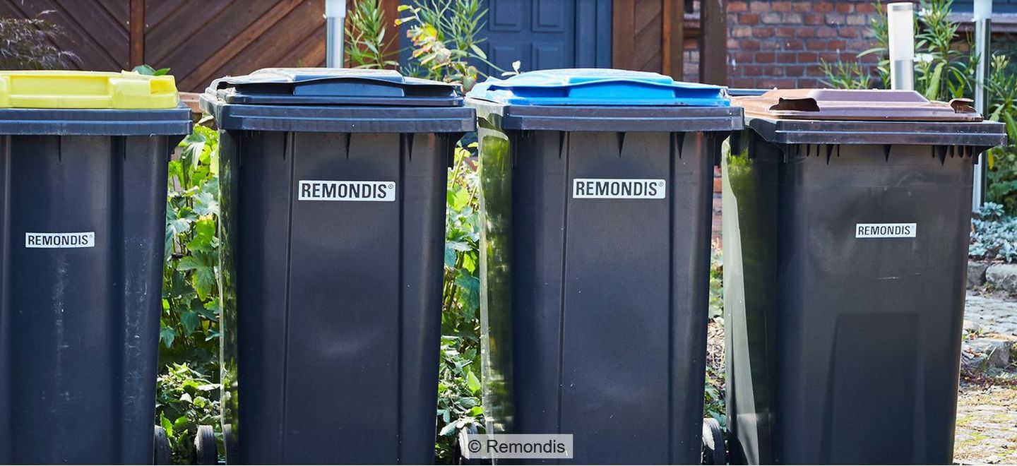 Vier Mülltonnen mit Aufdruck "Remondis" und verschieden farbigen Deckeln auf Fußweg, im Hintergrund Pflanzen, Hauswand und Eingangstür