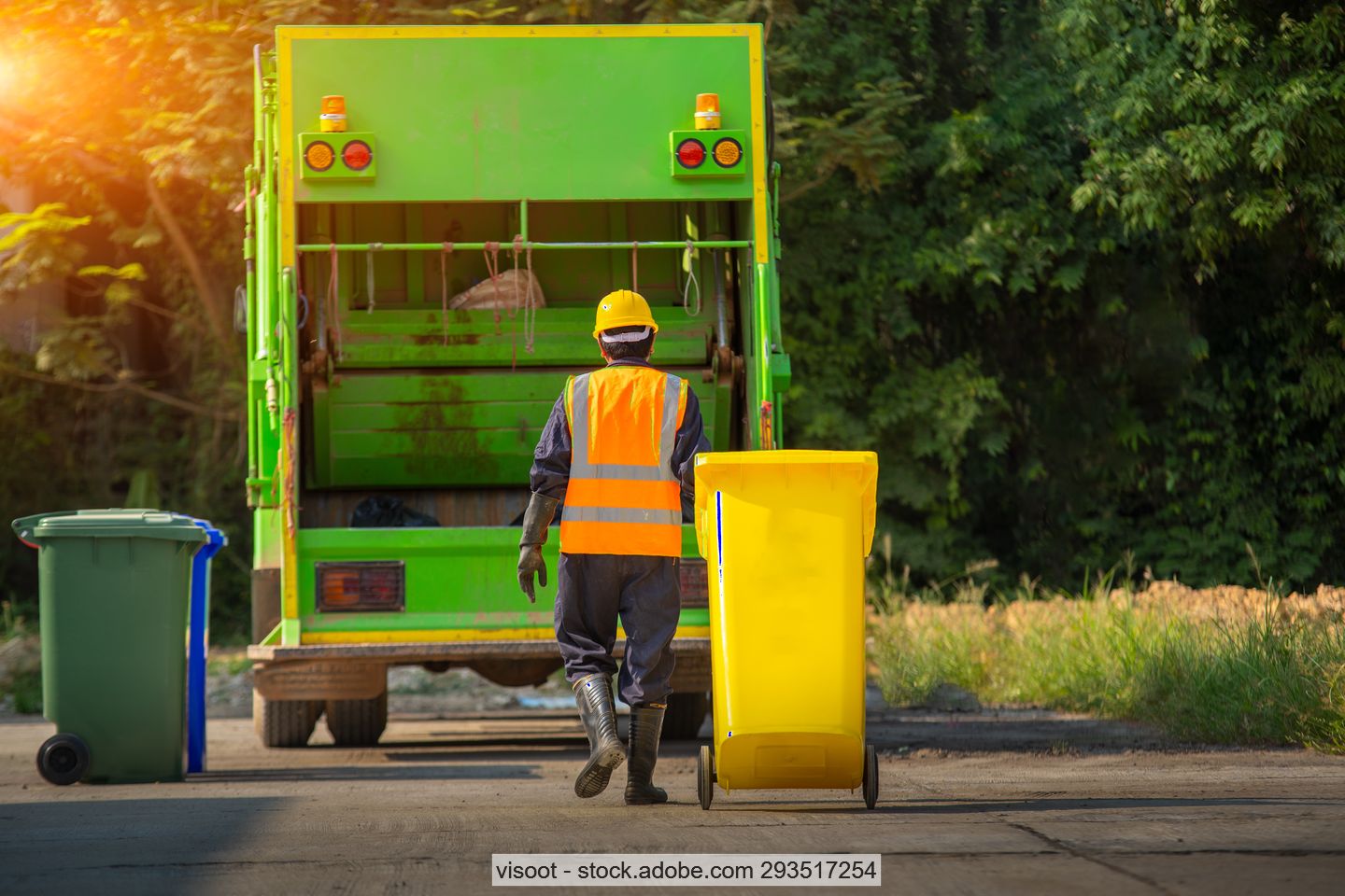 Müllwerker auf Straße mit gelber Abfalltonne hinter grünem Müllsammelfahrzeug, am Rand weitere Mülltonnen im Hintergrund Bäume