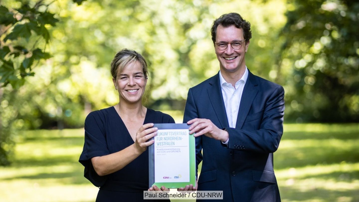 Mona Neubaur und Hendrik Wüst halten Exemplar eines gedruckten Koalitionsvertrages hoch im Hintergrund grüne Wiesen und Bäume